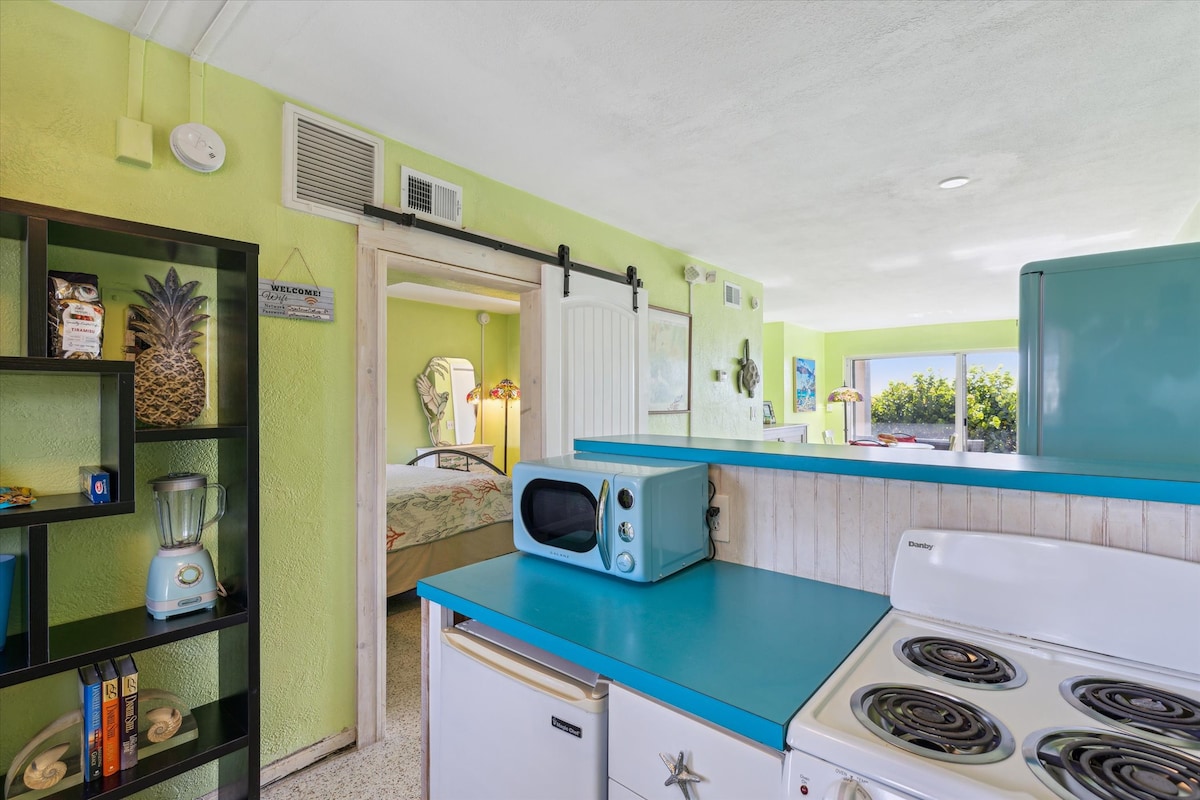 A compact kitchen space features a vibrant teal countertop with a range of appliances, including a stove, microwave, and blender. Light green walls add a fresh touch, while a doorway leads to a sleeping area, illuminated by natural light from a nearby window.