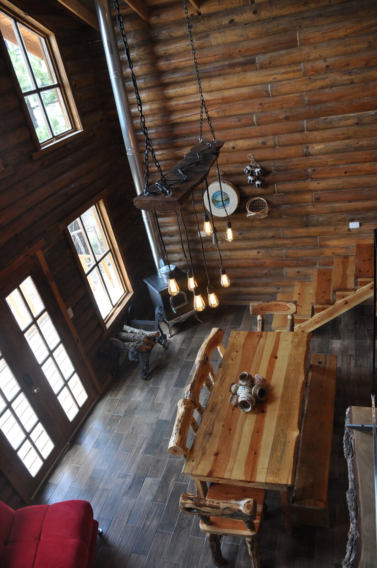 An overhead view of a rustic cabin interior showcases a wooden dining table surrounded by log-style chairs. A hanging light fixture with exposed bulbs illuminates the space. Large windows allow natural light to fill the area, enhancing the warm wood tones of the walls.
