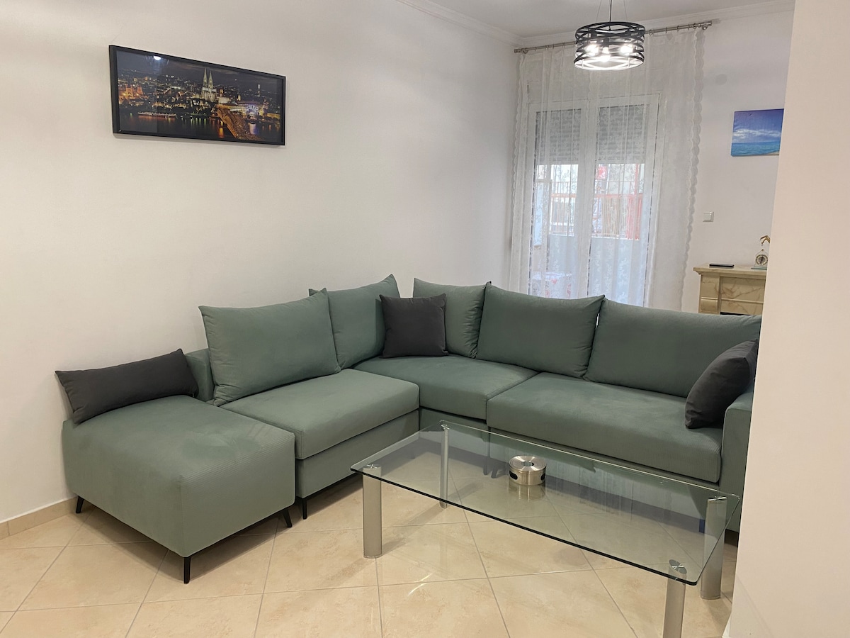 A modern living area features a large L-shaped sofa in muted green, accompanied by black accent pillows. A glass coffee table sits in front, reflecting soft natural light from a nearby window, which is dressed with sheer white curtains. A framed cityscape photograph adorns the wall.