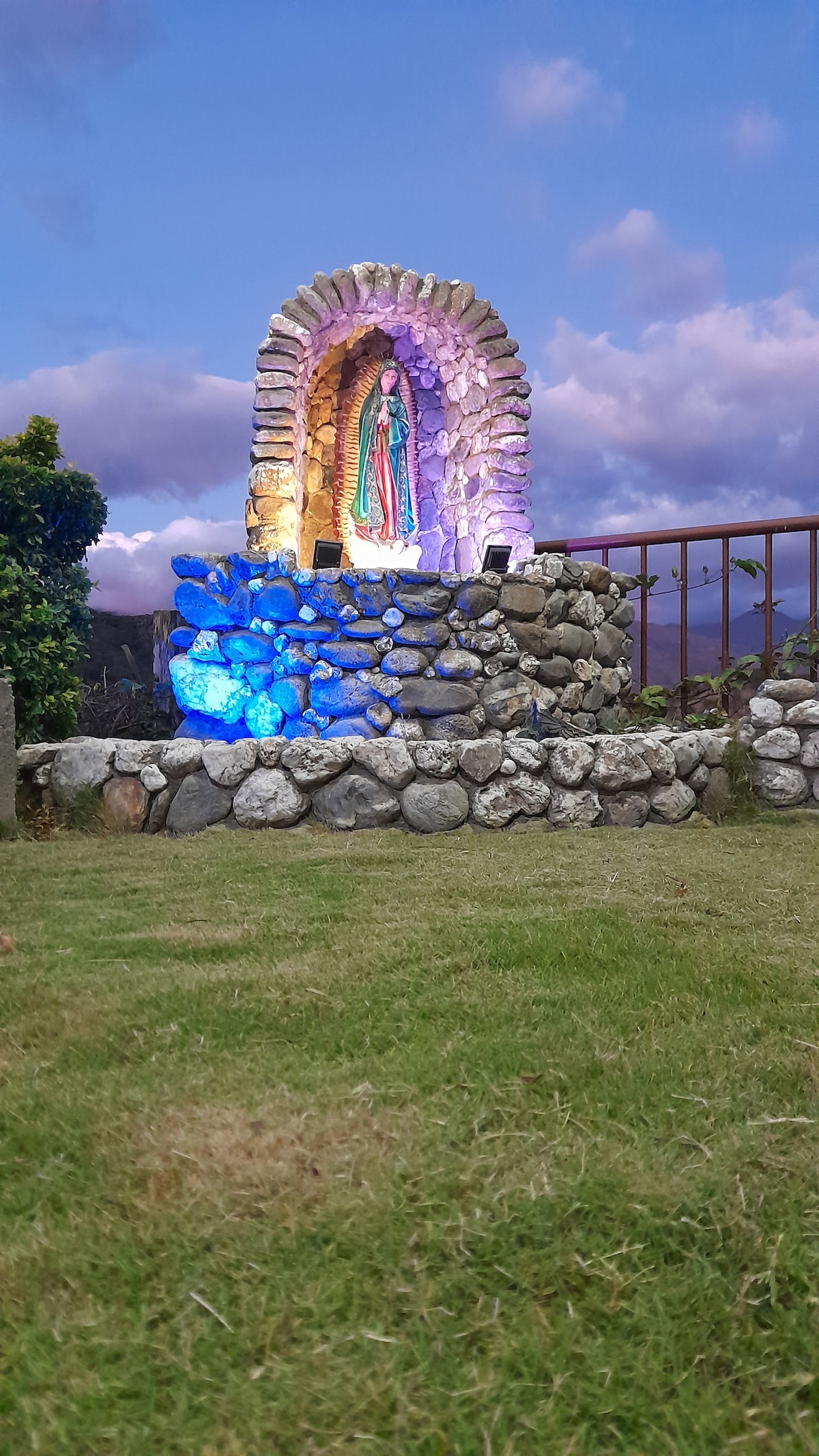 A beautifully crafted stone altar features a carved image of a figure, illuminated in shades of blue and purple. The altar is surrounded by smooth stones and set against a backdrop of green grass, with a scenic sky filled with soft clouds in the distance.