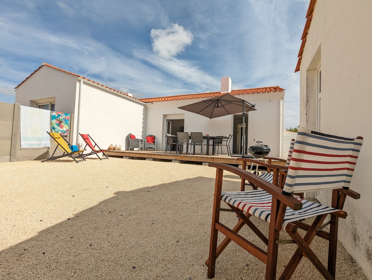 A patio area is featured with an outdoor dining set and shaded seating under an umbrella. Lounge chairs in bright colors provide additional relaxation spots, while a barbecue grill and a textured gravel surface complete the space, creating an inviting atmosphere for outdoor enjoyment.