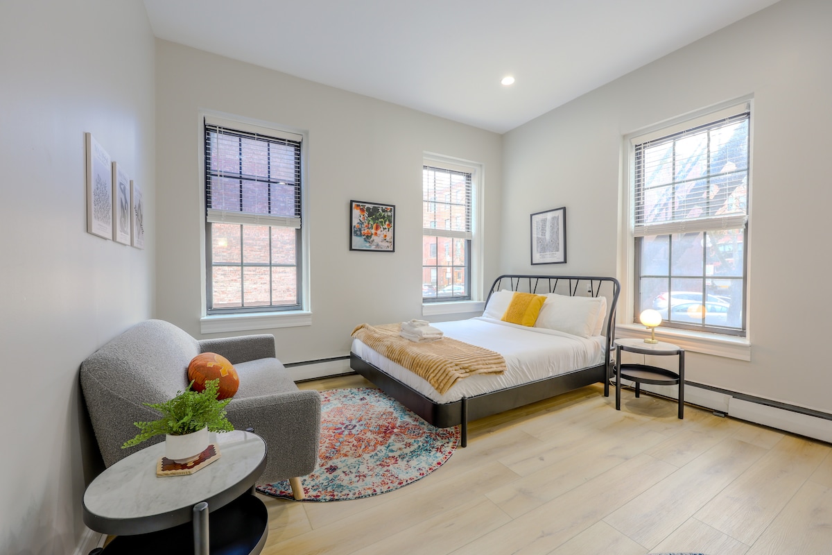 The studio features a spacious bed complemented by a soft throw blanket. Large windows allow natural light to fill the room, highlighting the light-colored wooden flooring. A small table with a plant and decorative globe sits beside a cozy seating area.