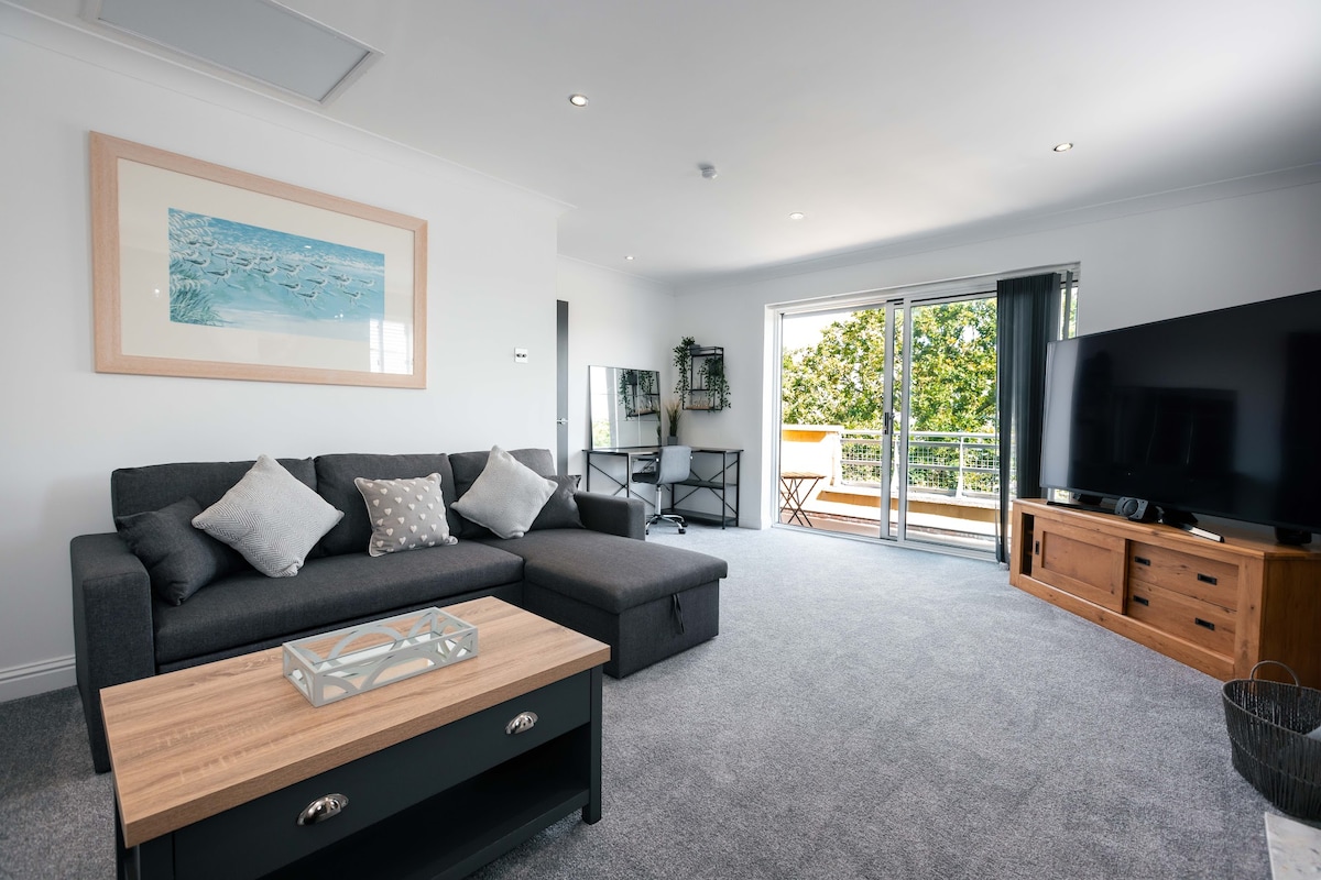 A spacious living room features a dark gray corner sofa with decorative cushions, a low wooden coffee table, and a large flat-screen TV on a wooden console. Glass doors open to a balcony, allowing natural light and views of the surrounding greenery.