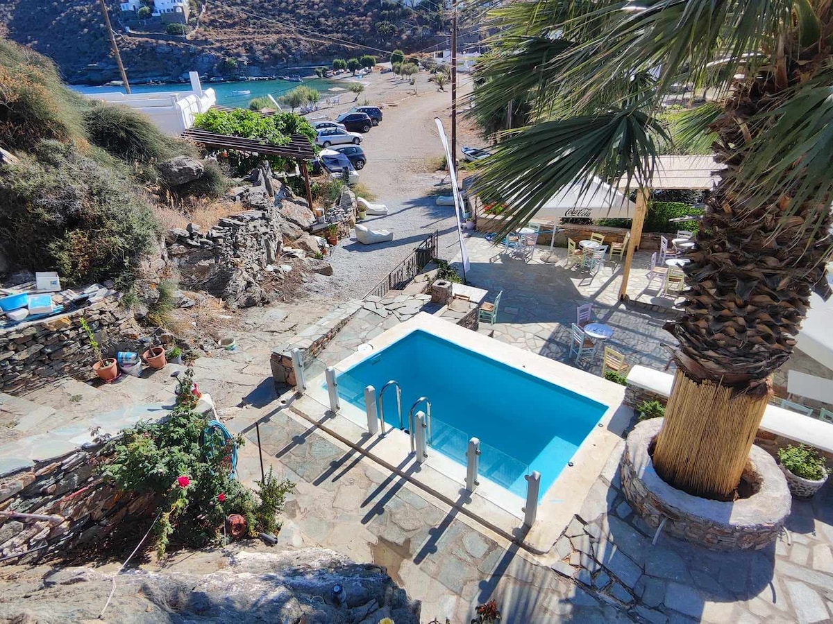 A private swimming pool is set in a stone-paved outdoor area, surrounded by lush greenery and colorful flowers. Sun loungers are positioned nearby, with a view of the landscape and distant shoreline visible beyond.