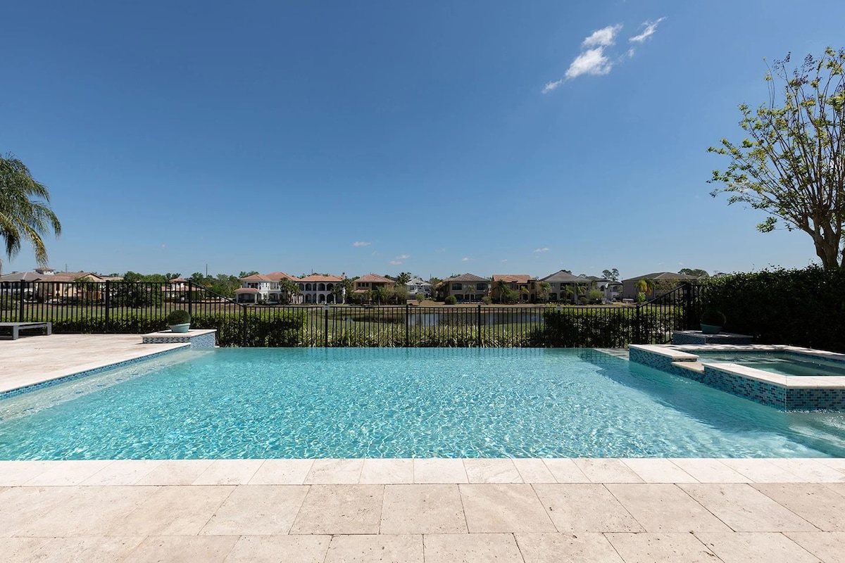 A spacious infinity pool sparkles under clear blue skies, bordered by light stone tiles. Views extend over green grass and residential buildings in the distance, while a separate spa is visible adjacent to the pool, creating a tranquil outdoor retreat.