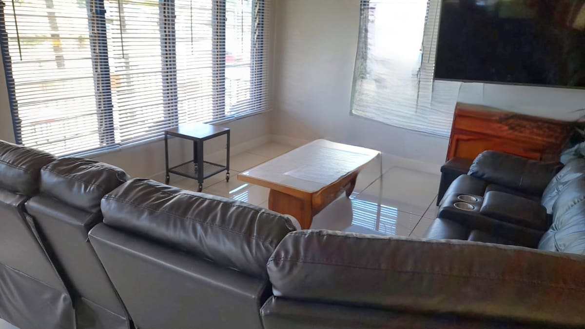 A cozy living room is featured, showcasing a spacious leather sectional sofa oriented towards a mounted television. A wooden coffee table sits centrally, while large windows with blinds allow natural light to fill the space. The room is designed for relaxation and casual gathering.