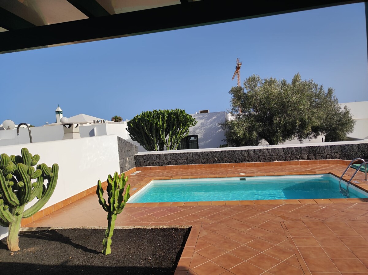 A refreshing swimming pool is nestled within a private outdoor area, framed by various desert plants and cacti. The tiled patio extends around the pool, providing ample space for relaxation. Light blue water contrasts with the terracotta flooring under a clear blue sky.