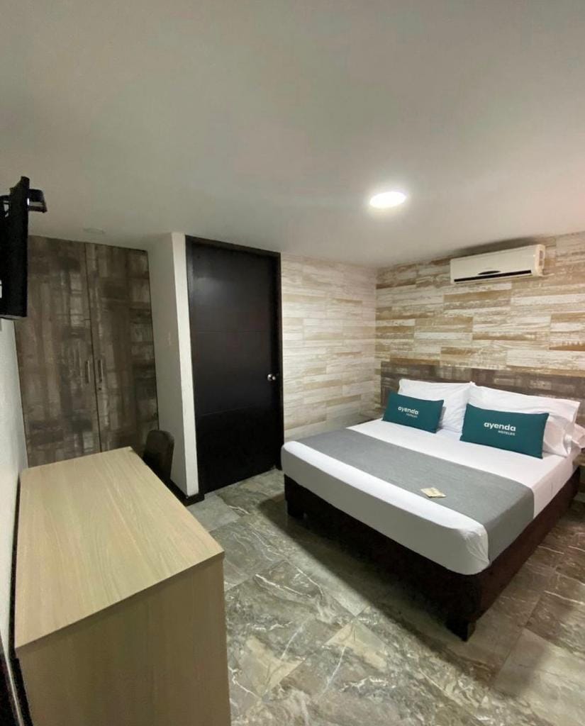 A comfortable room features a spacious bed with a gray and white bedding ensemble. An air conditioning unit is above the bed, and a closet offers storage. A flat-screen TV is mounted on the wall. Natural light is provided by the ceiling fixture.