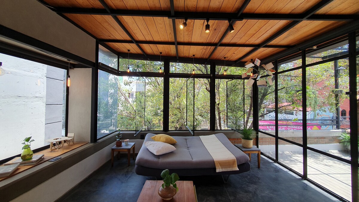 A bright and airy room is surrounded by large glass windows, offering views of trees outside. A spacious bed is situated in the center, complemented by wooden beams on the ceiling. Natural light streams in, enhancing the soothing atmosphere of the space.
