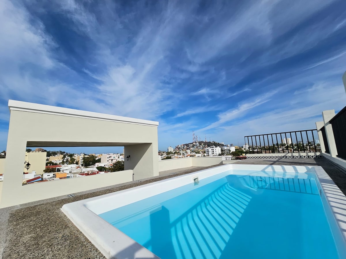 A rooftop pool reflects bright sky blues, offering a wide view of the surrounding neighborhood and distant hills. The pool area is bordered by a sleek railing, and sunlight creates playful patterns on the water's surface.
