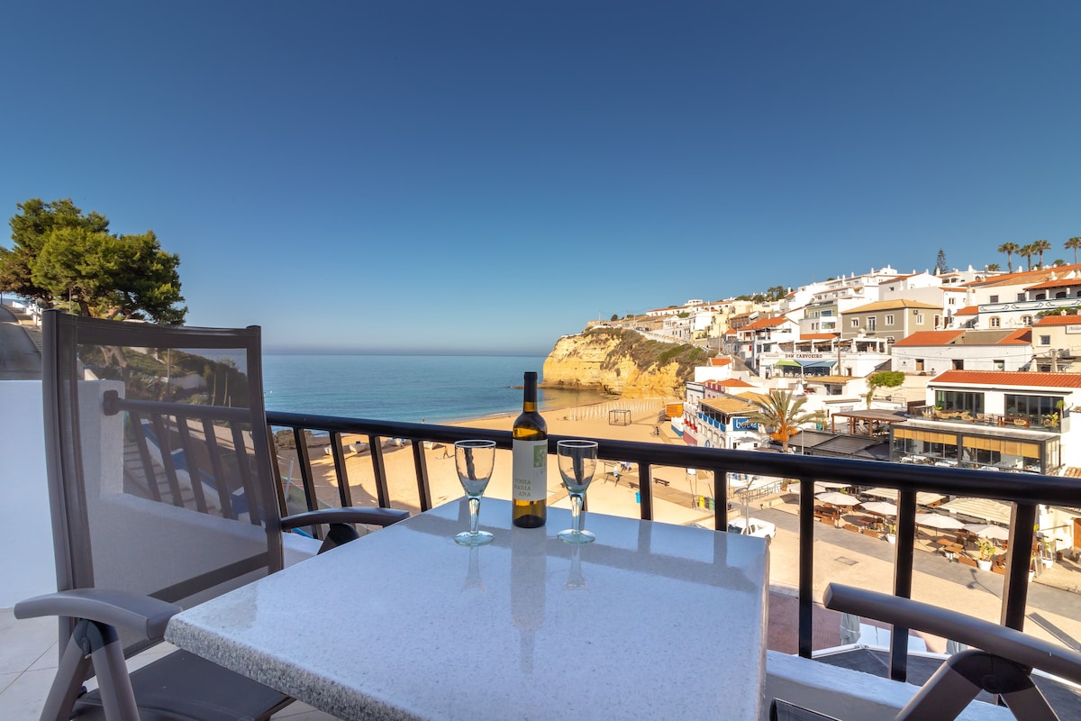 A balcony table is set for two, featuring a bottle of wine and two glasses. The view overlooks a serene beach and coastline, providing a glimpse of surrounding buildings and palm trees. The clear blue sky complements the tranquil ocean scene.