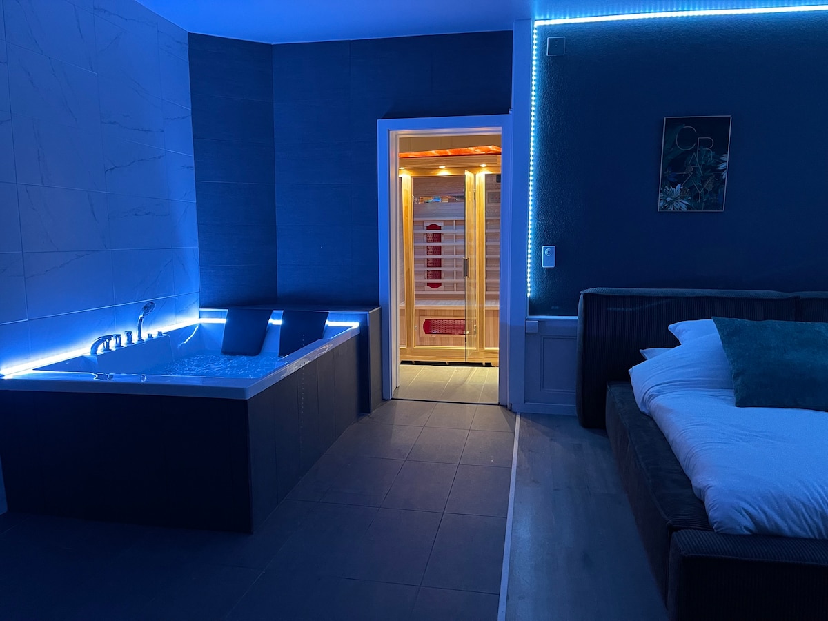A spacious suite interior features a luxurious jacuzzi tub illuminated by soft blue lighting, adjacent to a sauna with a glass door. A cozy bed with neutral bedding is positioned nearby, enhancing the tranquil atmosphere of relaxation.