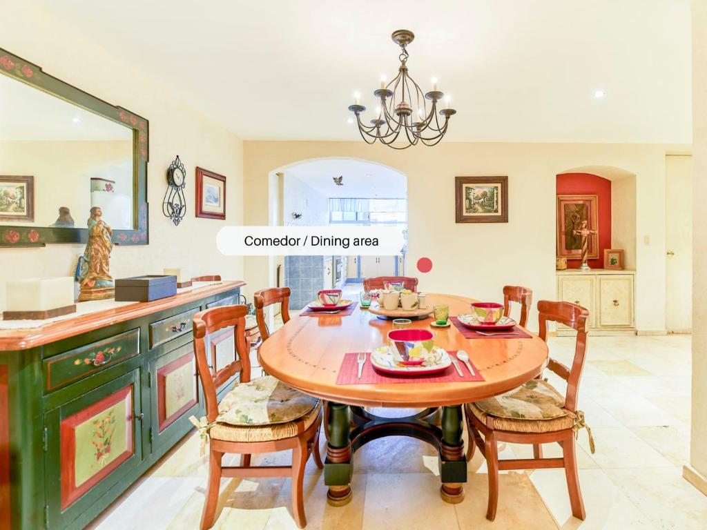 A dining area features a large wooden table surrounded by several chairs, each adorned with cushions. The table is set with colorful plates and utensils, while a chandelier hangs above. Decorative items and artwork enhance the warm ambiance of the space.