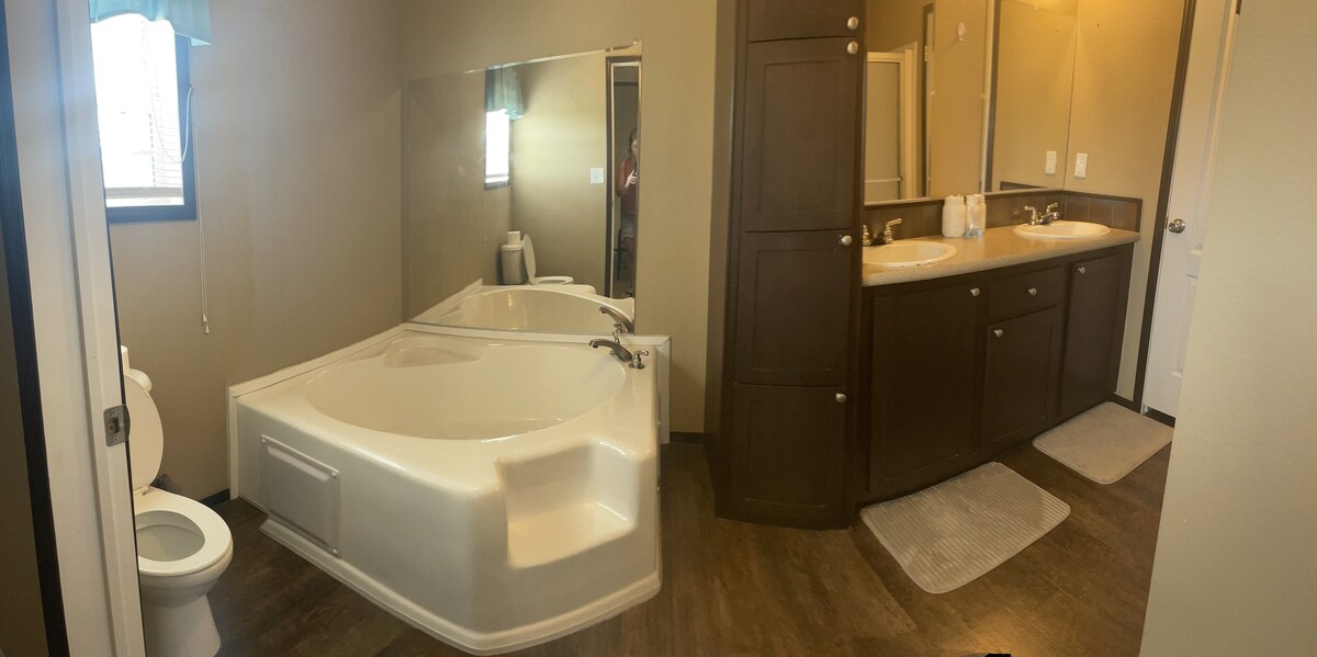 A spacious bathroom is featured with a large, white soaking tub and dual vanities. Light fixtures above the sinks illuminate the area, while a mirror reflects the room's features. A neutral color palette creates a calm ambiance, complemented by wooden cabinets and flooring.