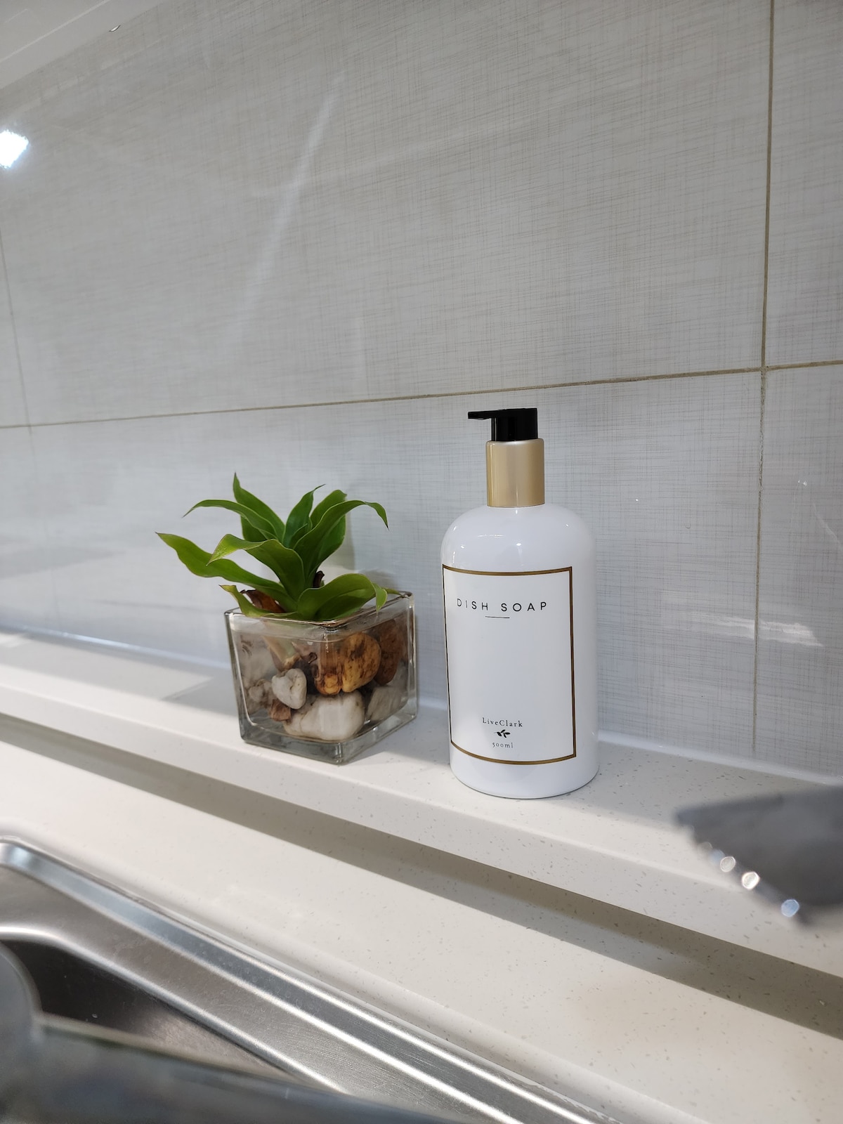 A modern kitchen sink area features a tall bottle of dish soap and a decorative glass container filled with natural elements. A small green plant adds a pop of color, complementing the clean and contemporary design of the space.