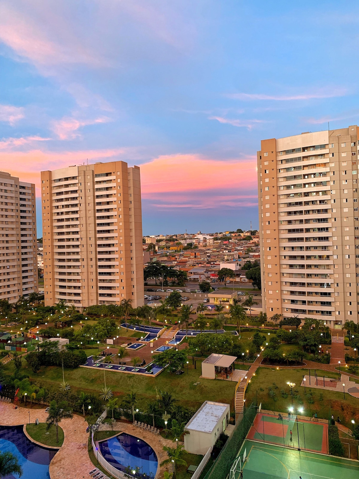 Two tall buildings are positioned on either side of the image, overlooking landscaped gardens and recreational areas below. A vibrant sky transitions from blue to warm pink hues as sunset approaches, providing a serene backdrop to the setting.