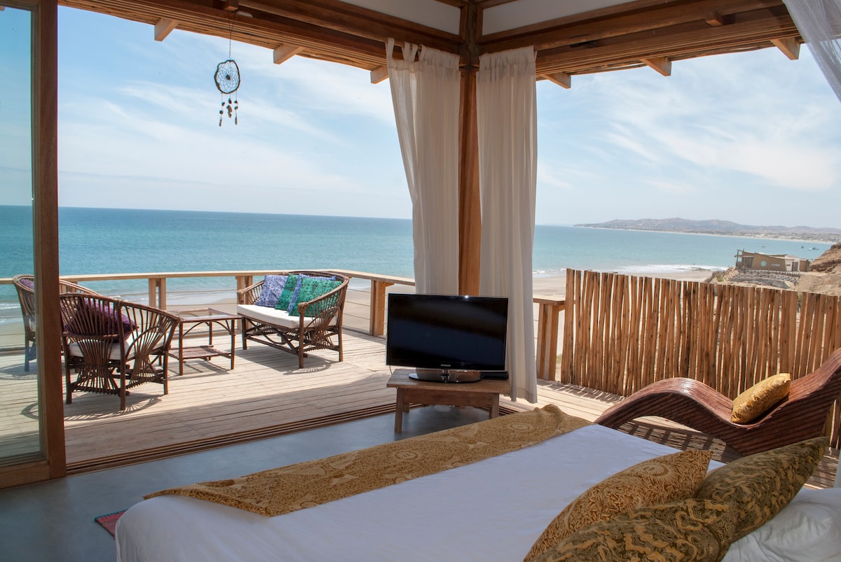 A spacious bedroom is presented with a large bed facing expansive glass doors that open to a terrace overlooking the ocean. A cozy seating area is seen outside, complemented by woven chairs and vibrant throw pillows. Natural light floods the room, creating a bright and airy atmosphere.