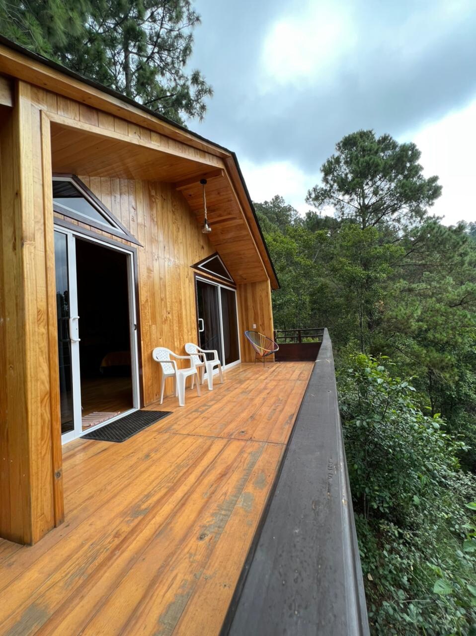 The exterior of a wooden cabin is highlighted, showcasing a spacious deck with two white chairs. Large glass doors open to the interior, framed by natural wood, providing views of the surrounding trees and lush greenery.