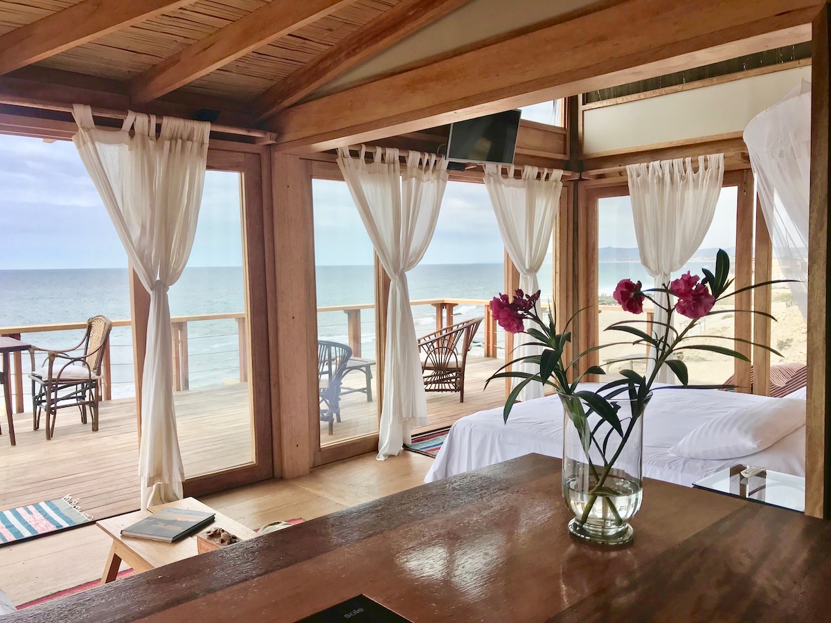 A spacious living area opens to breathtaking ocean views through large glass doors. Light soft curtains frame the windows, and a simple vase holds vibrant flowers on a wooden table. Comfortable seating arrangements can be seen on the deck outside, merging indoor and outdoor spaces.