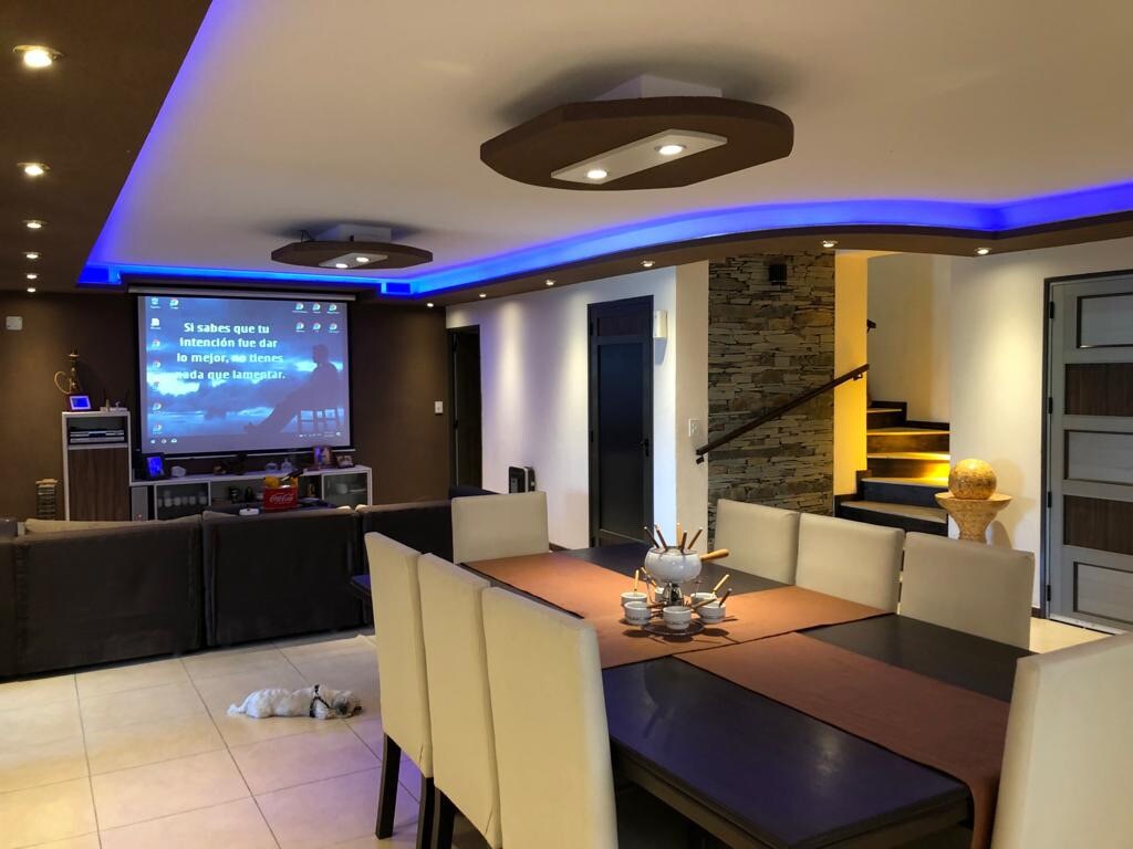 A spacious dining area is showcased with a large rectangular table surrounded by light-colored chairs. Soft blue ambient lighting highlights the room. A projection screen is mounted on a wall, while modern ceiling fixtures provide additional illumination. Stairs lead to an upper level in the background.