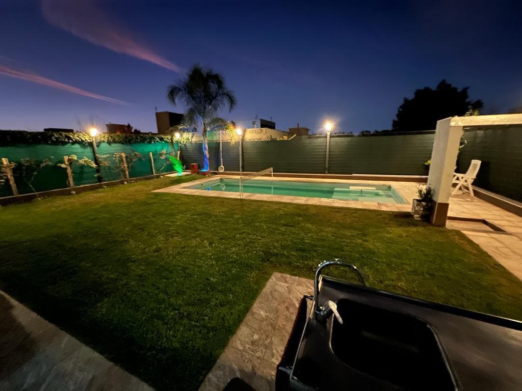An outdoor area features a well-maintained grassy lawn surrounding a swimming pool, softly illuminated under evening lights. Palm trees and decorative plants are positioned along the perimeter, creating a pleasant atmosphere. A patio area includes seating for relaxation.