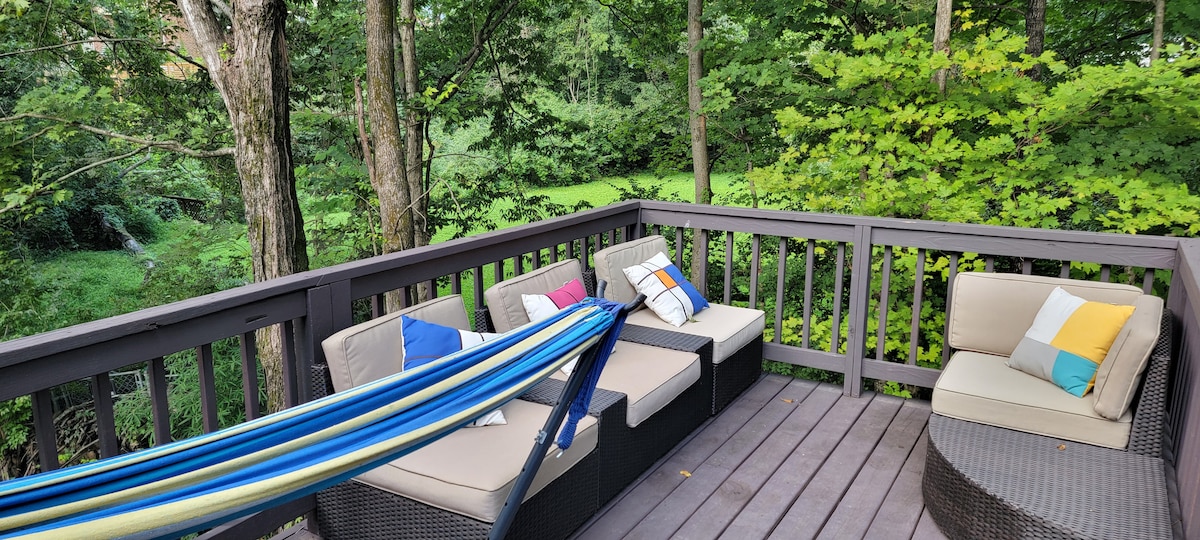 A shaded back patio is shown, featuring comfortable seating arrangements with colorful cushions. A hammock is suspended between two posts, inviting relaxation, while greenery and trees create a serene, natural backdrop, enhancing the peaceful outdoor environment.
