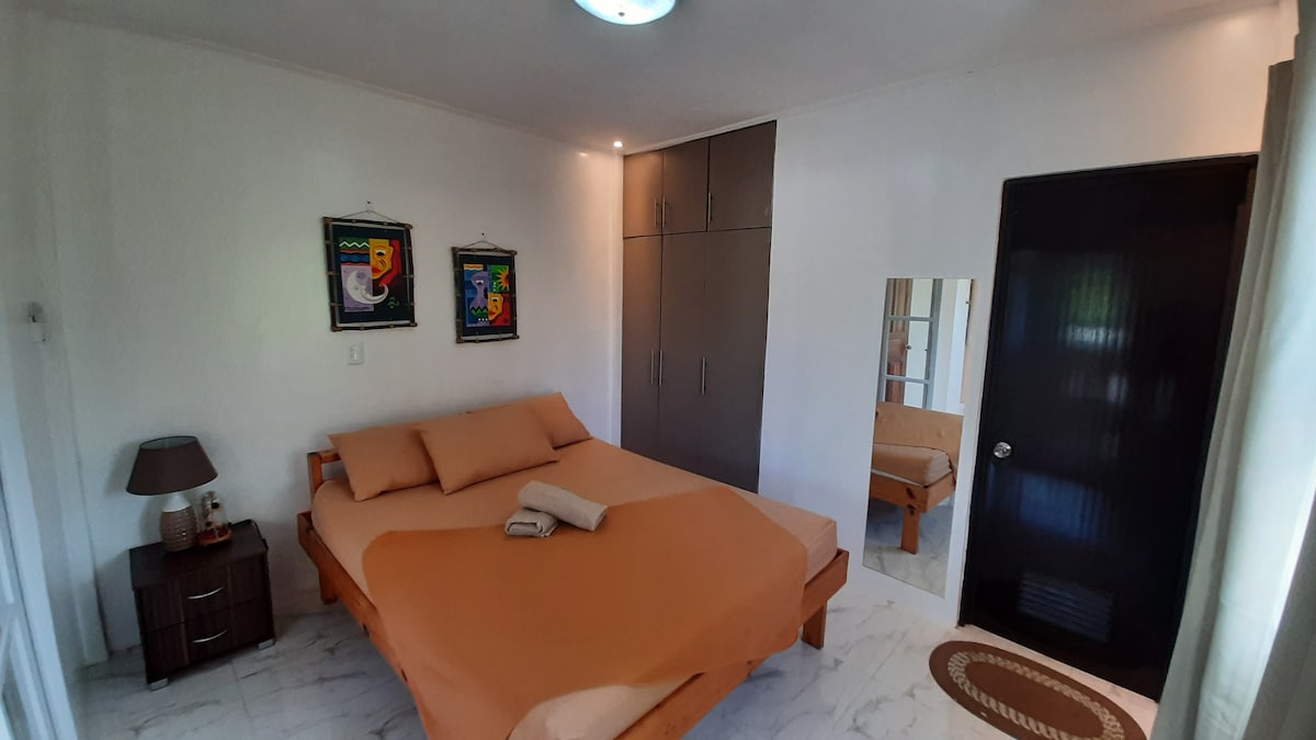 A spacious sleeping area is highlighted by a large bed with a neutral-colored duvet and two pillows. It features bedside tables with lamps, framed artwork on the walls, and a wardrobe. Soft natural light enters through a nearby window, enhancing the inviting atmosphere.