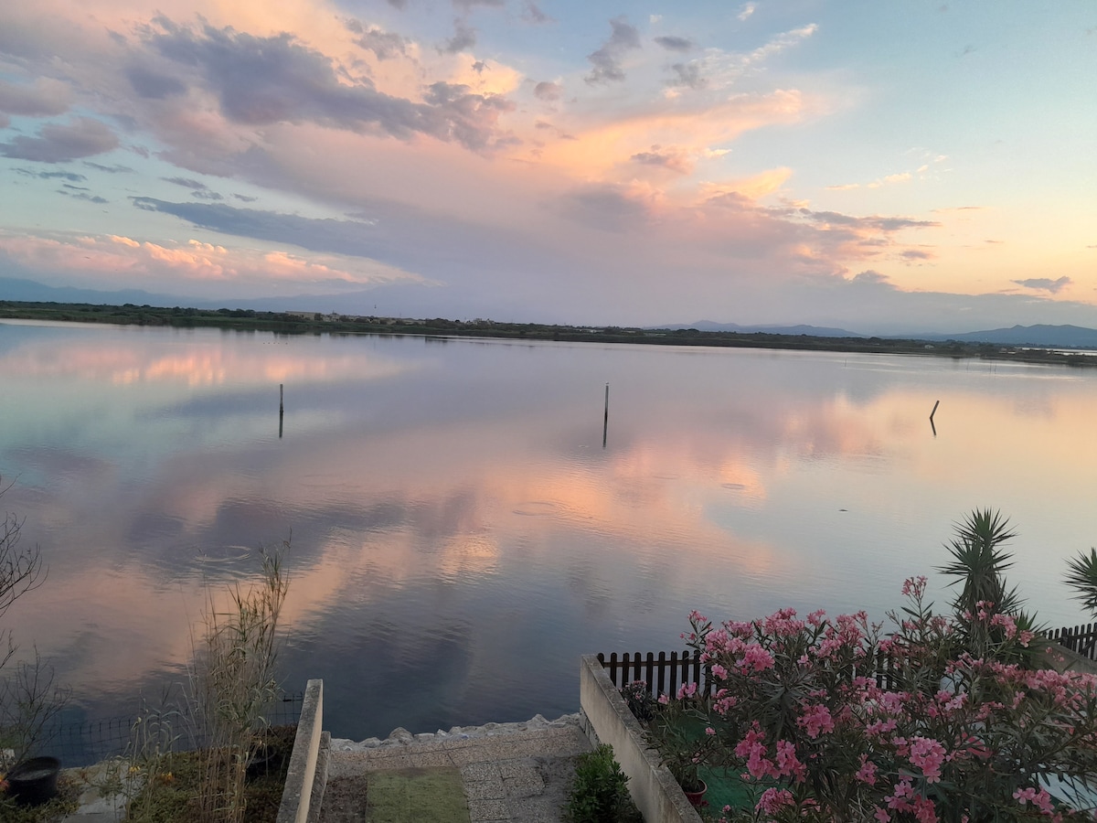 A tranquil image captures a water landscape reflecting soft hues of pink and blue from the sky above. Silhouettes of distant mountains are visible, while pink flowers and greenery frame the foreground, leading to the calm water's edge.