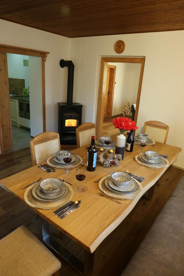 A dining table is set with elegant tableware, including plates and glassware, arranged on woven placemats. A bottle of wine and a candle are positioned centrally, while a warm glow is cast by a nearby wood stove. The room is surrounded by wooden accents.
