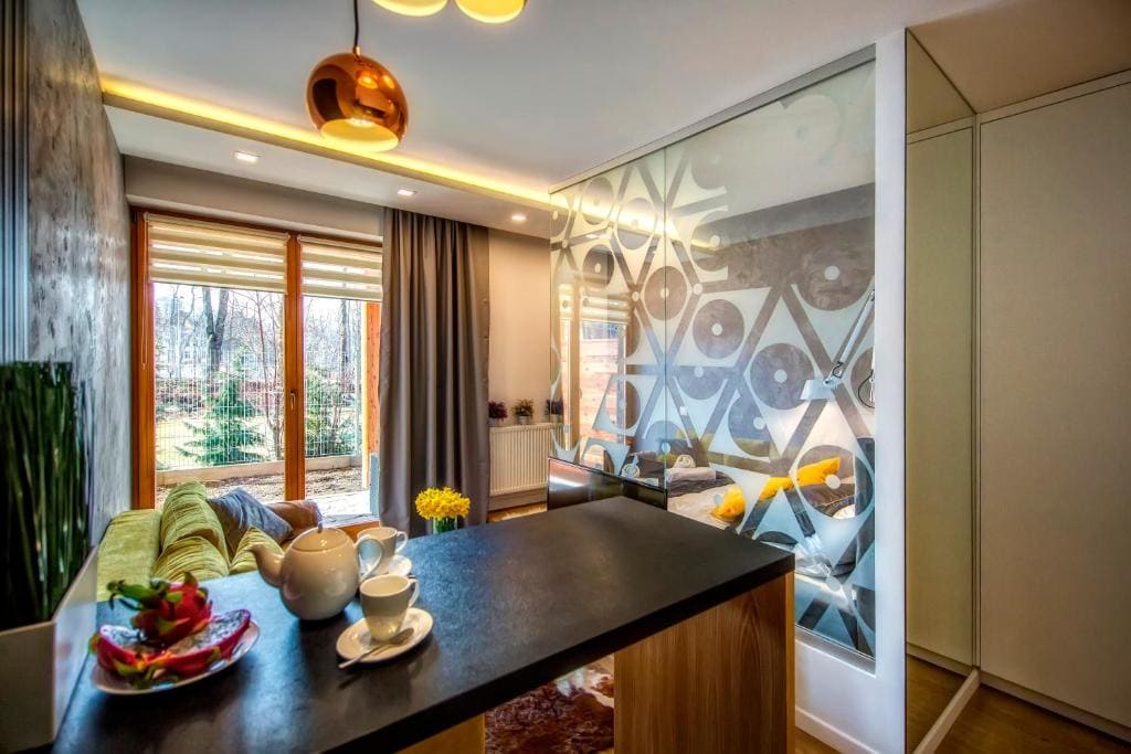 A modern living space features a small kitchenette with a countertop. A bowl of fruit and two cups are positioned on the counter. A green sofa provides seating, while large glass doors allow natural light to fill the room, opening to a view of greenery outside.