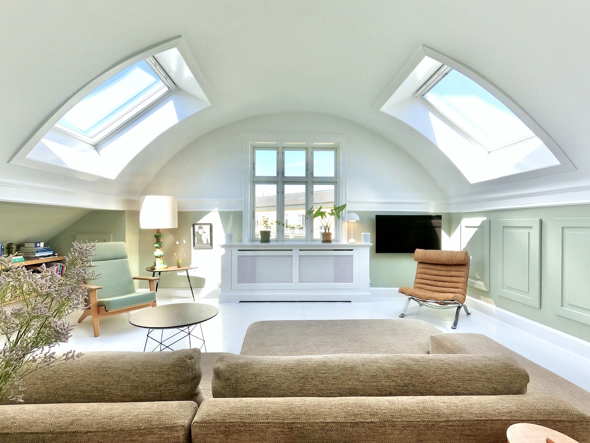 A bright living space features sloping ceilings adorned with skylights allowing ample natural light. A comfortable seating area is arranged with a soft sofa and a modern chair, complemented by a minimalistic coffee table. A serene palette of soft green and white enhances the overall ambiance.