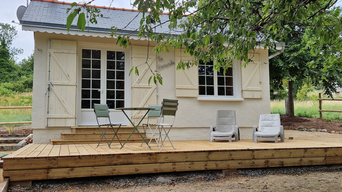 A welcoming exterior view of a single-story home showcases a wooden deck with a small round table and two chairs, complemented by two additional lounge seats. Light-colored shutters frame the windows, providing a charming accent against the neutral wall tone.