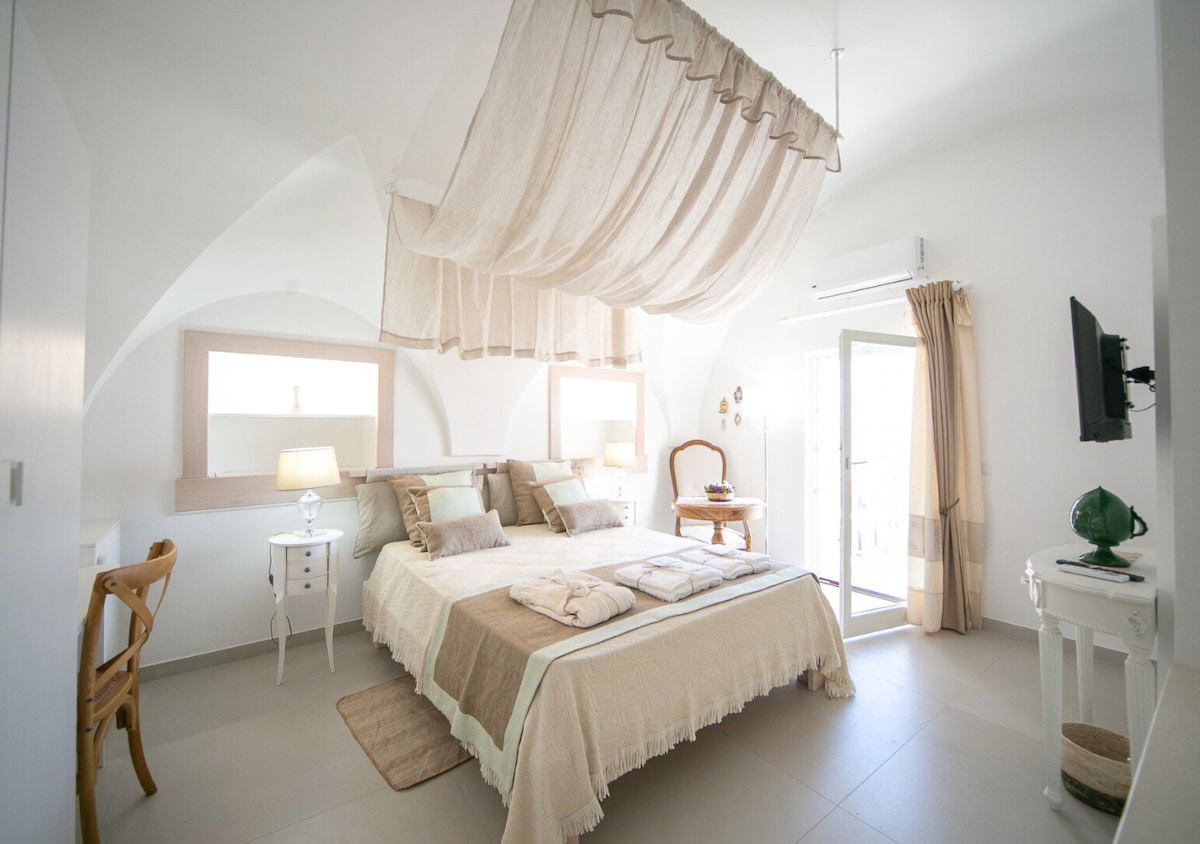 A spacious sleeping area is highlighted by soft, neutral tones and a gently draped fabric over the bed. Two bedside lamps provide subtle lighting, complemented by natural light from windows. A chair and a television are also present, enhancing the room's functionality.