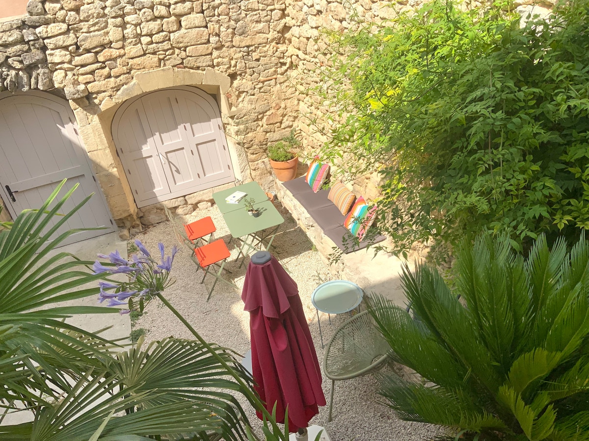 A charming courtyard is framed by a stone wall, featuring a small table and two red chairs. A long bench with colorful cushions rests against the wall, surrounded by lush greenery. A large umbrella provides shade, adding to the relaxing atmosphere.
