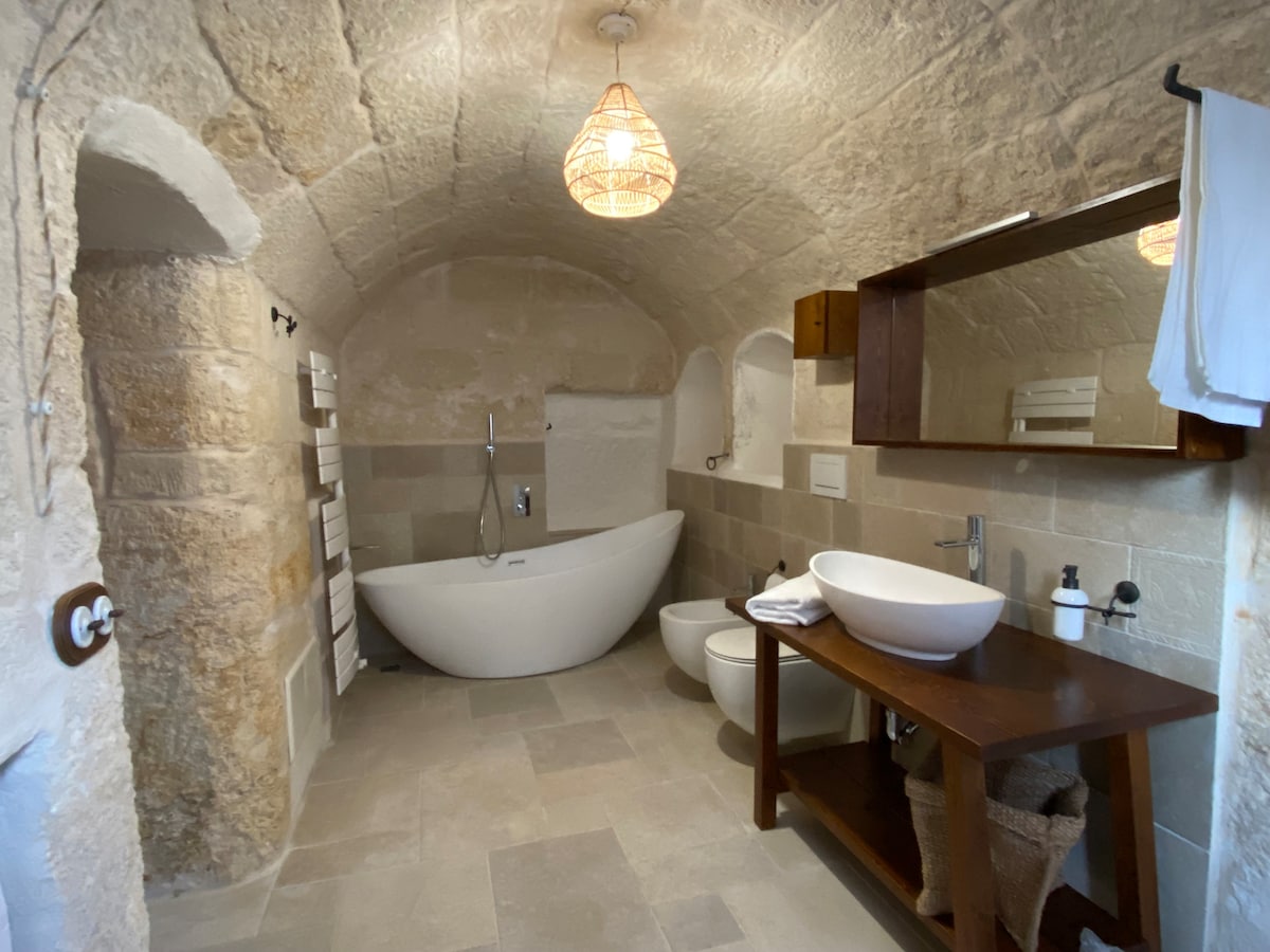 A spacious bathroom showcases a freestanding bathtub and a modern toilet. Natural stone walls and a ceiling create a rustic ambiance. A wooden vanity holds a sink and accessories, while a mirror reflects light. Soft lighting is provided by a pendant fixture.