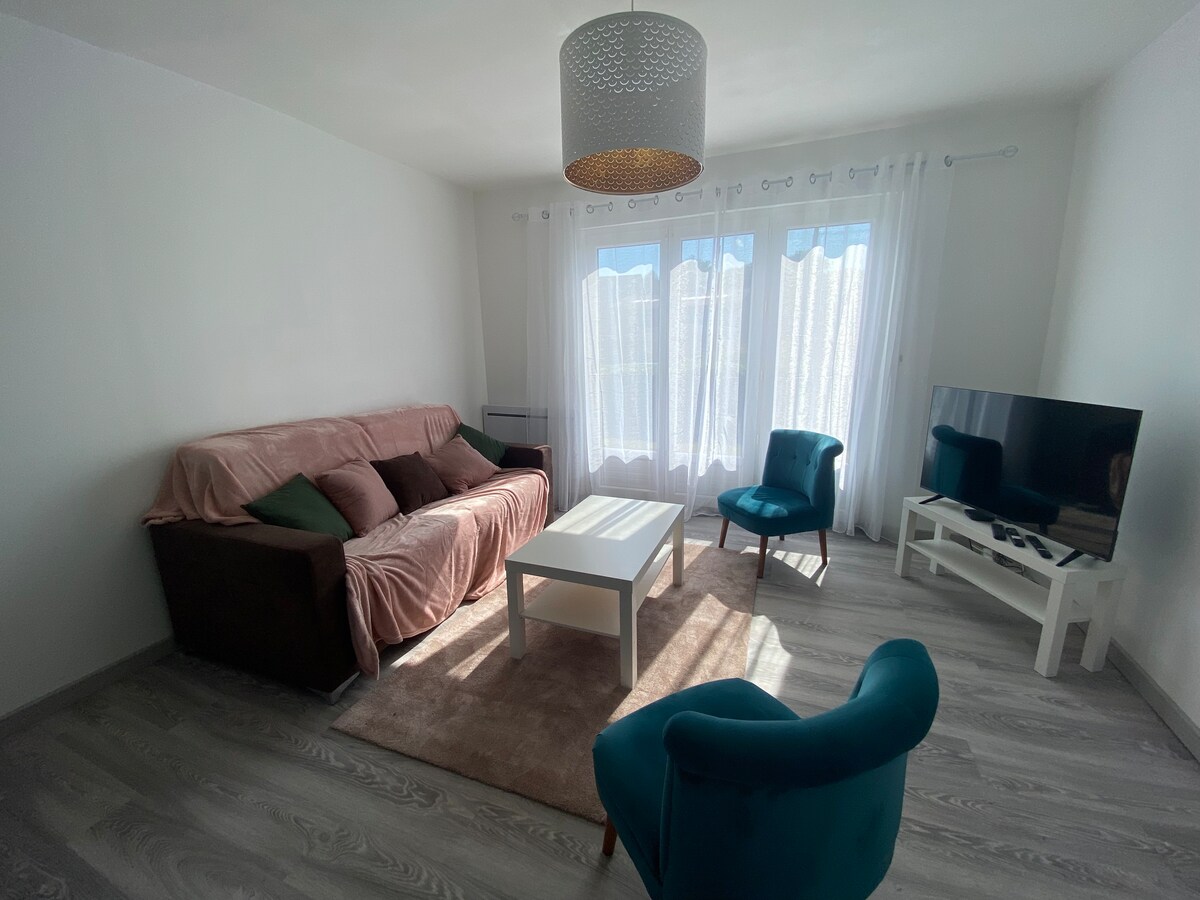 A bright living area features a comfortable sofa covered with a soft throw and decorative cushions. A coffee table sits in the center, complemented by a light blue armchair. Large windows are dressed in sheer drapes, allowing natural light to illuminate the space.