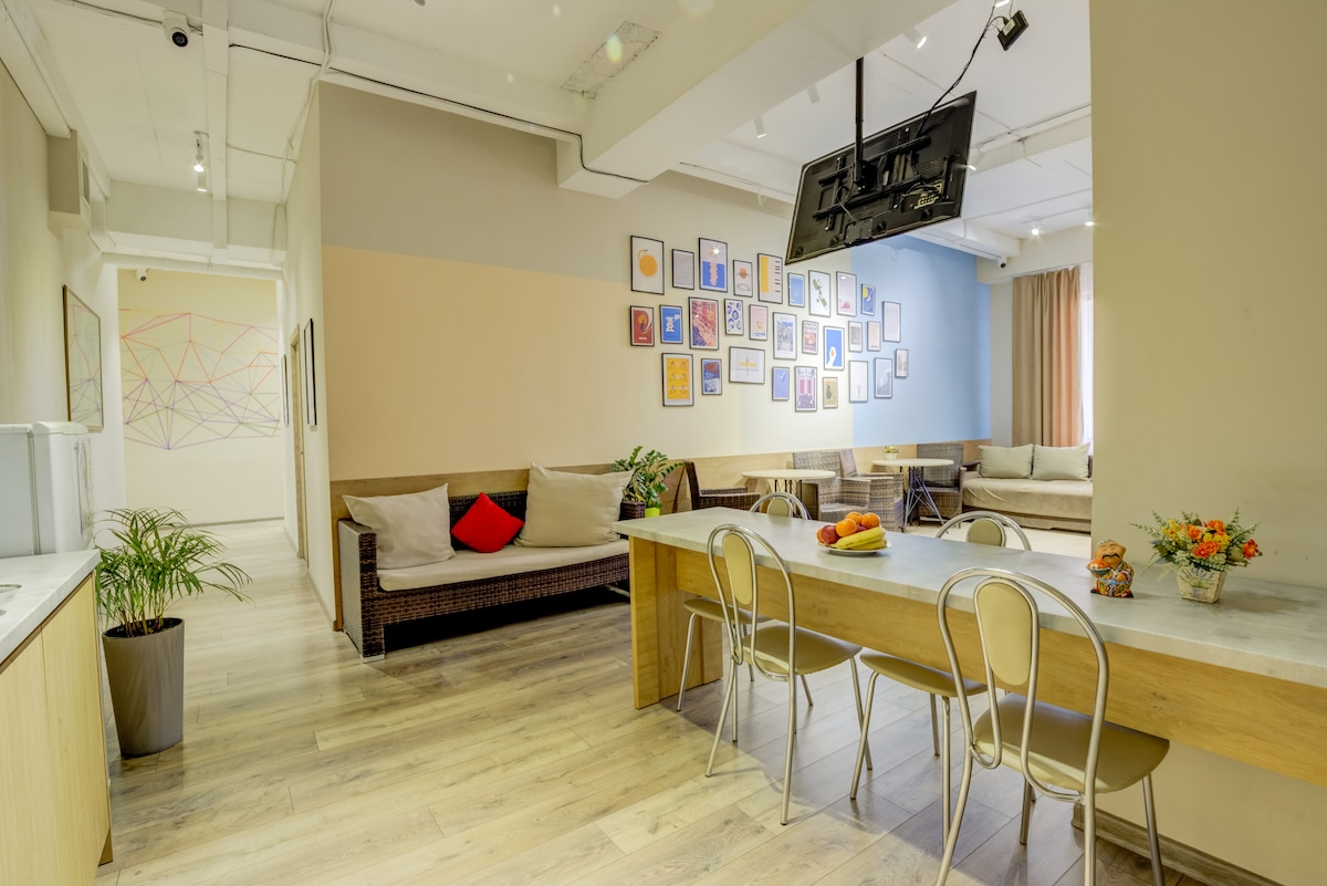 An inviting common area features a long dining table with modern chairs, surrounded by a stylish sofa arrangement. Colorful artwork adorns the wall, while a vibrant flower arrangement and fresh fruit are displayed on the table, adding character to the space.