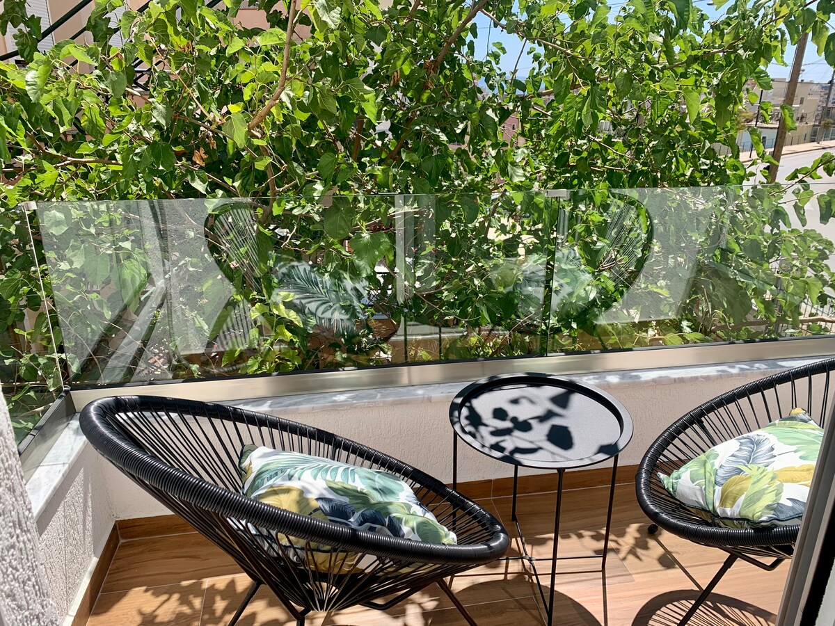 A private balcony area is furnished with two black woven chairs featuring colorful cushions, accompanied by a small round table. Lush greenery is visible through a glass barrier, providing a serene outdoor space.