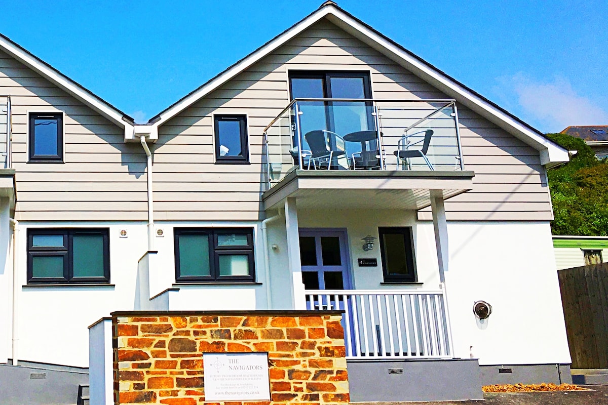 The exterior of a contemporary townhouse features a light grey and white facade with multiple windows. A balcony with glass railing is visible above the entrance, furnished with two chairs and a small table. A brick wall stands at the front, adding texture to the appearance.