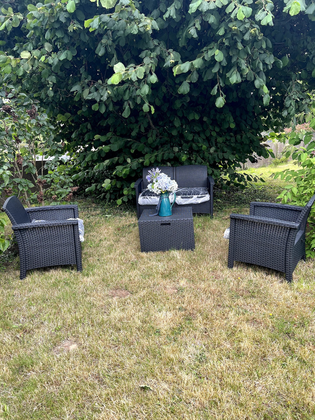 A garden seating area is surrounded by lush greenery, featuring four black wicker chairs arranged around a small square table. A vase of flowers is placed on the table, adding a touch of color to the outdoor space. The area is set on a grassy lawn.