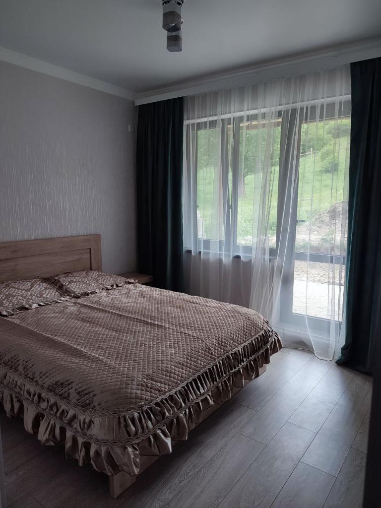 A sleeping area features a spacious bed dressed in a textured brown quilt with a ruffled edge. Large windows allow natural light to filter through sheer curtains, offering a view of the greenery outside. Smooth wooden flooring complements the neutral wall colors.