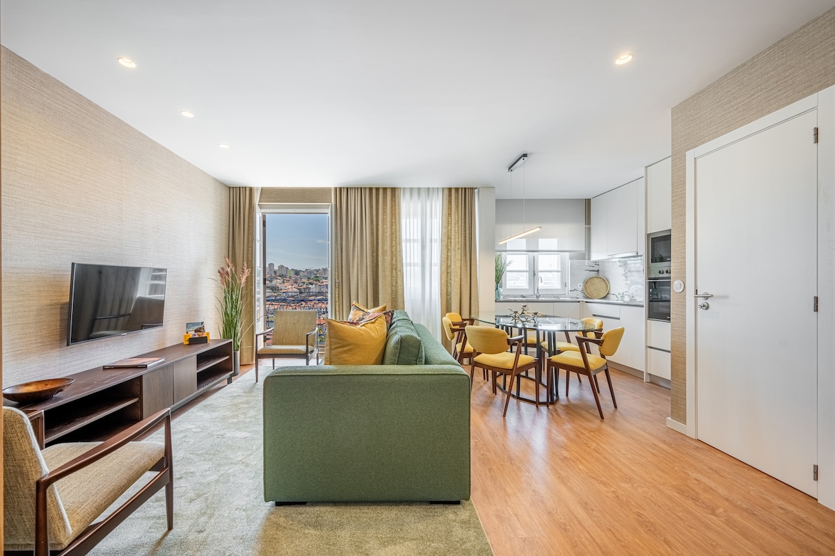 A modern living area features a green sofa and complementary armchairs arranged around a wooden entertainment unit and a coffee table. A dining table with chairs is visible, along with a well-equipped kitchen. Natural light enters through large windows, offering a city view.