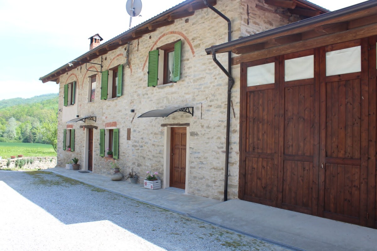 A rustic stone building with green shutters is depicted, complemented by wooden accents. The entrance is framed by a simple awning, and a large garage door is visible. The surrounding landscape features lush greenery, creating a serene rural setting.