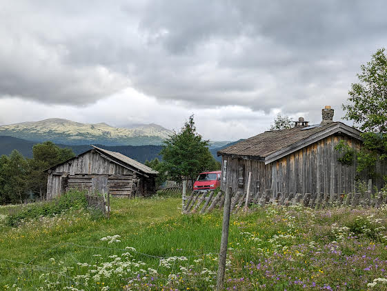 Two rustic wooden buildings are set against a backdrop of rolling hills and mountains. Lush green grass and a variety of wildflowers surround the structures, with a hint of distant peaks visible under a cloudy sky. A red vehicle is parked nearby.