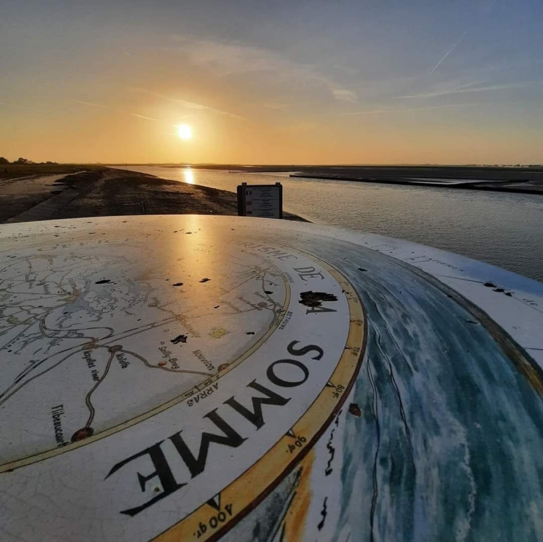 A scenic view capturing the sun setting over the Baie de Somme, with a map featuring the region prominently displayed in the foreground. The tranquil water reflects the soft glow of the sunset, while the shoreline gently curves into the distance.