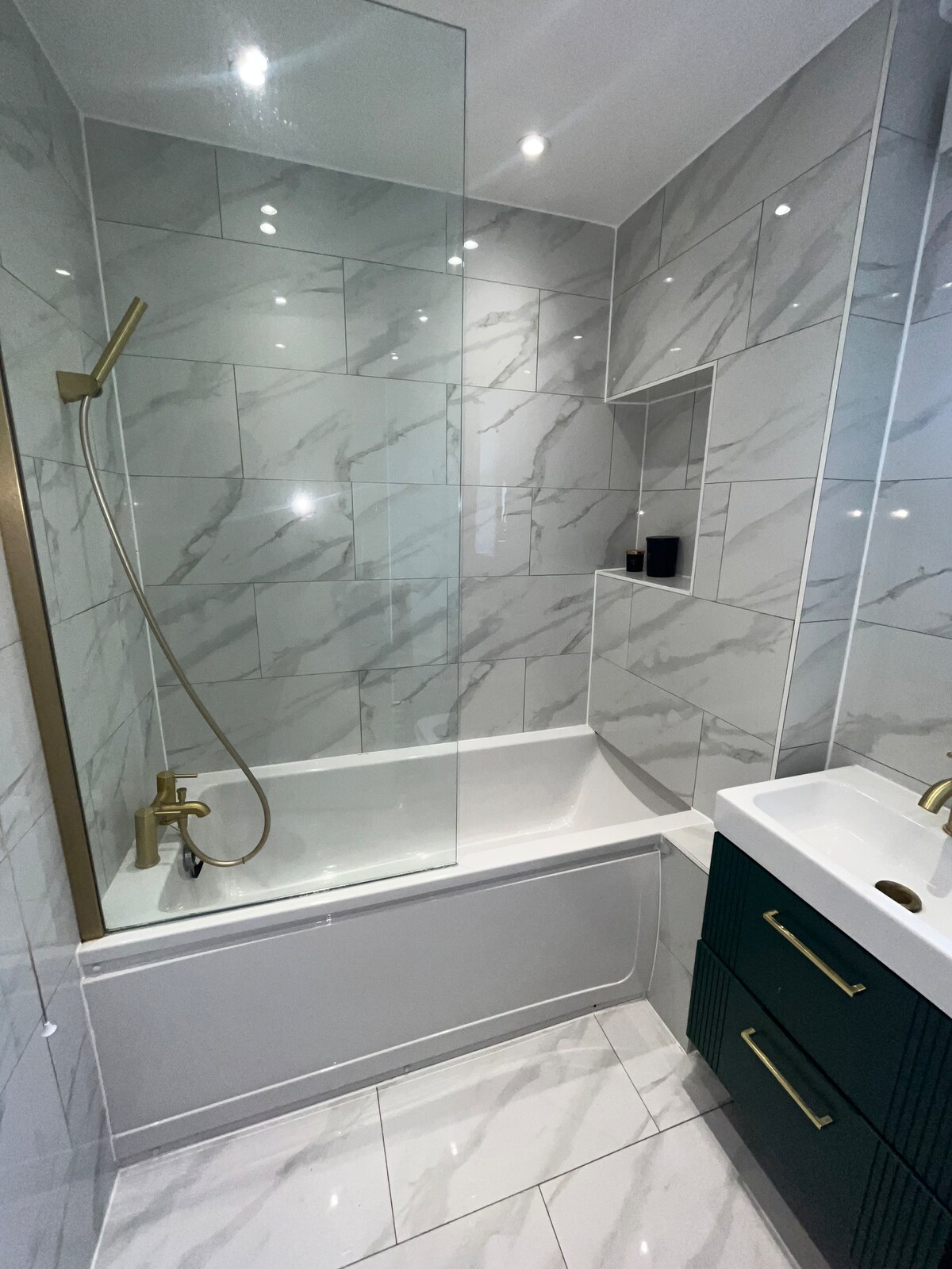 A modern bathroom features elegant marble tiles on the walls and floor. A glass shower enclosure is positioned above a bathtub, with a stylish brass showerhead. A vanity with dark green drawers and a white sink is present, illuminated by recessed lighting.
