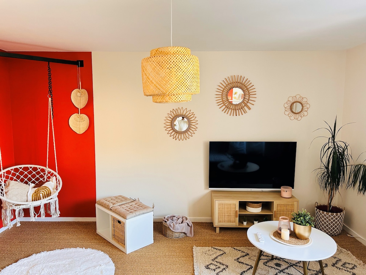 A cozy living area features a hanging chair, adding a relaxed element to the space. A television is positioned on a light wood stand, surrounded by decorative mirrors. Soft lighting is provided by a woven pendant lamp, complementing the warm tones of the wall.