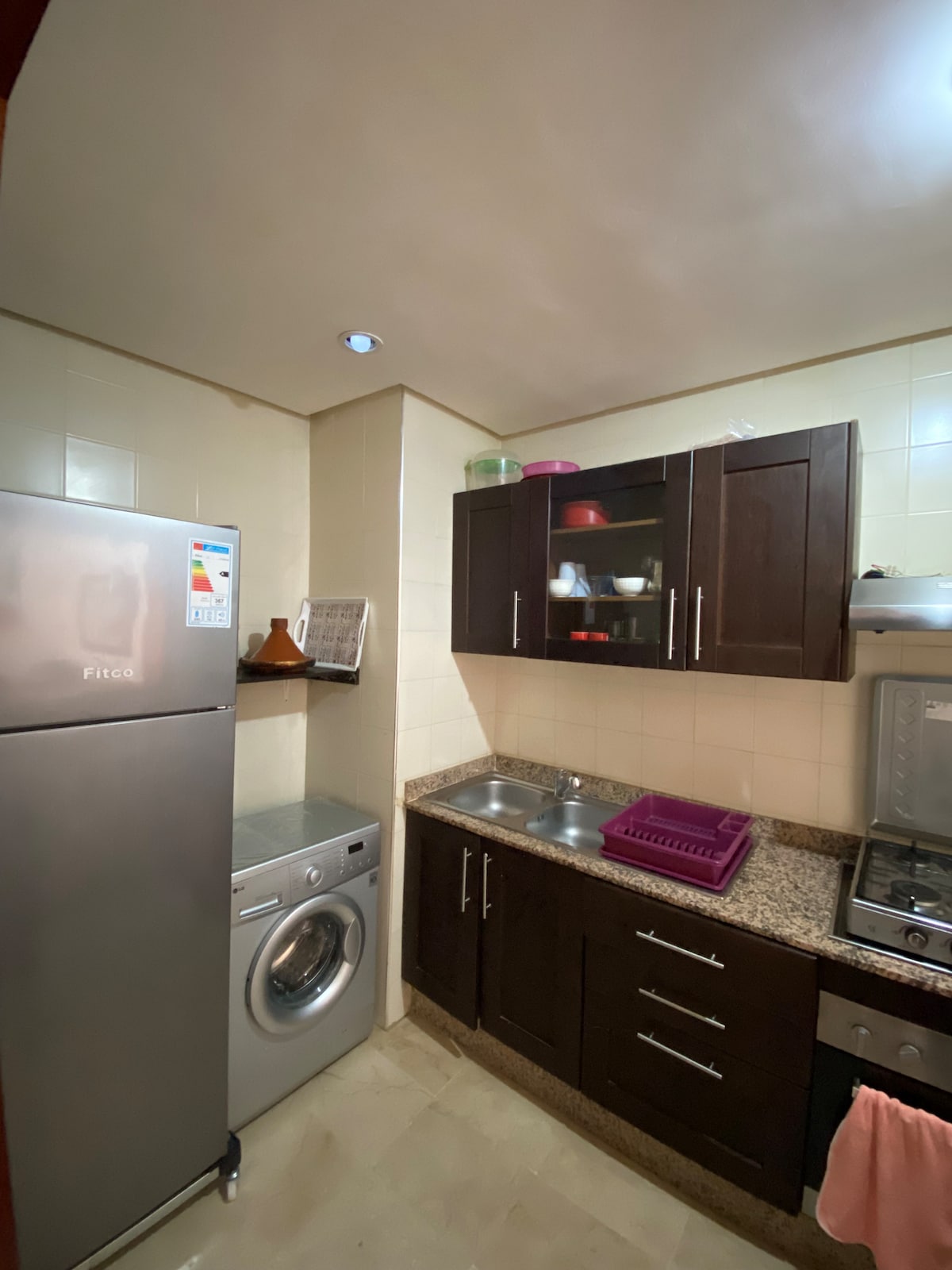 The kitchen features modern cabinetry with a dark wood finish. Stainless steel appliances, including a refrigerator and a washing machine, are visible. A granite countertop offers ample preparation space, alongside dual sinks and a gas stove. Bright overhead lighting enhances the functional layout.
