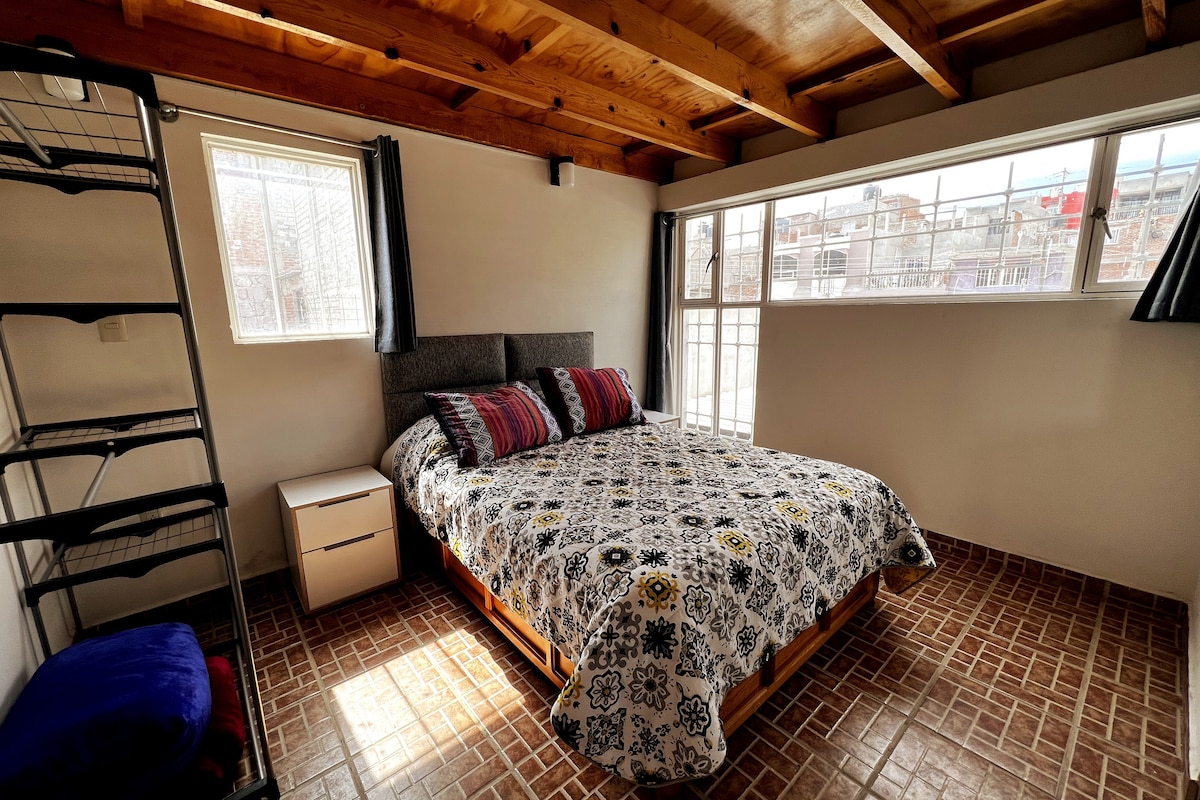 A bright sleeping area features a spacious bed adorned with a patterned blanket. Natural light fills the room through two windows, with a cozy nightstand nearby. A shelving unit stands against the wall, providing additional storage space.