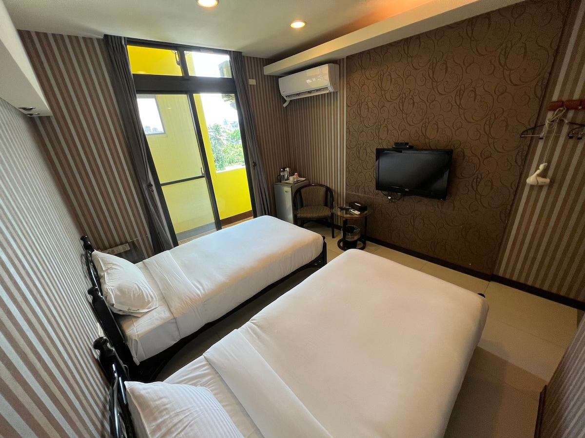 The room features two single beds positioned along the walls, with a television mounted on one side. A small chair is located near a window that opens to a balcony, allowing natural light to fill the space. Neutral tones and subtle patterns decorate the walls.